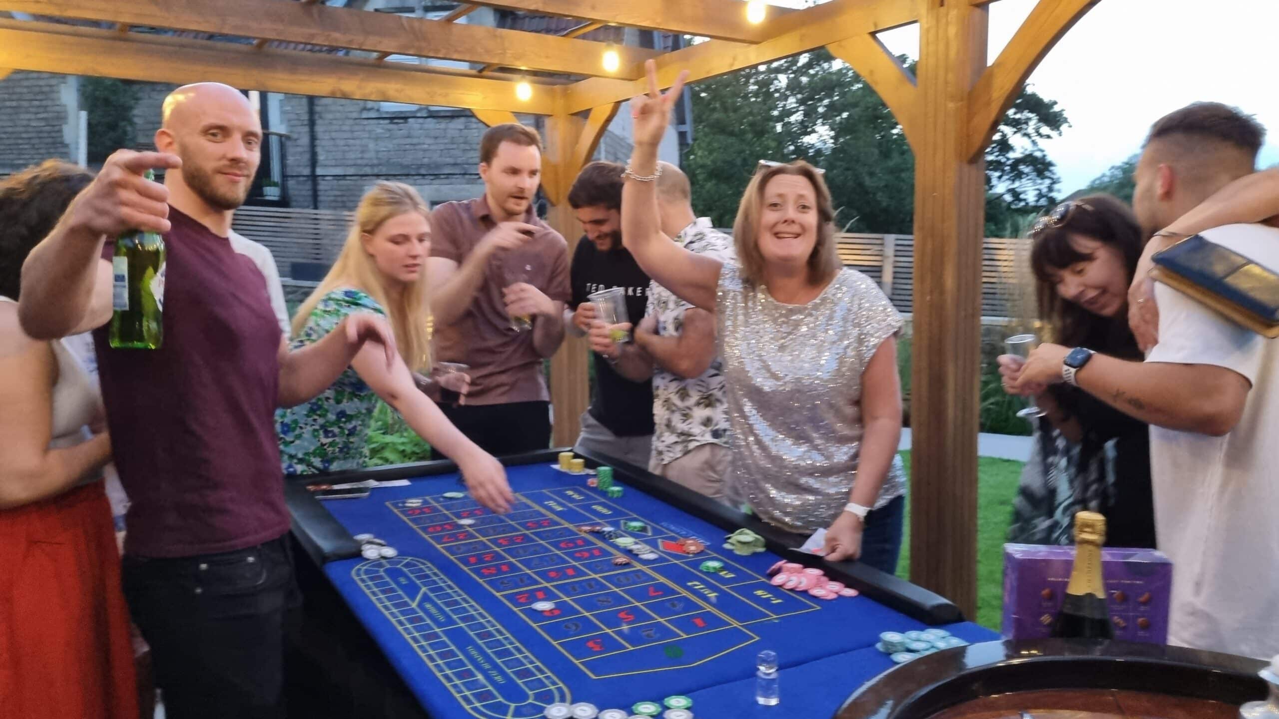 Party guests celebrating around a fun casino roulette table in a garden under a wooden pergola